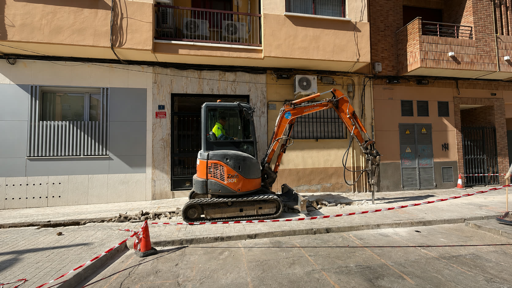 Miniexcavadora con martillo hidráulico levantando pavimento junto a una fachada